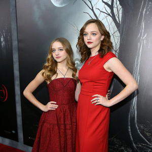 Two young actresses, Emily Banks and Fiona Dourif, posing dramatically at the 'Curse of Chucky' premiere. Dark, eerie backdrop with shadows and red accents.
