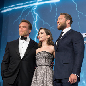Arnold Schwarzenegger, Emilia Clarke, and Jai Courtney on stage. Attending European premiere of Terminator Genisys. Wearing formal outfits. Large blue background with lightning graphic
