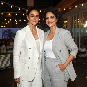 Gul Panag and Isha Narang at a book launch event. They are wearing matching white outfits and gray blazers. The backdrop is an indoor venue with lights and fans in the background.