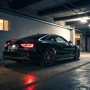 A shiny black Audi S5 coupe parked in a garage. It's spotlighted with dramatic lighting, showing off its sleek design and unique tail lights