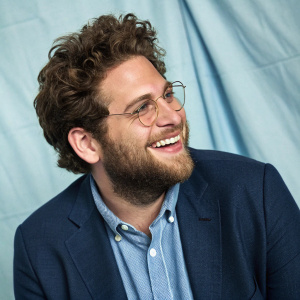 Jonah Hill, with a scruffy beard and brown curls, in a casual blue shirt and blazer. Smiling for a candid photo shoot. Studio backdrop with light blue fabric.