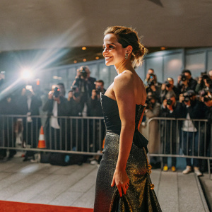 Emma Watson, at the New York Film Festival premiere of 'Beauty and the Beast'. Dressed in a black and gold sequined gown. Crowds of photographers and fans in the background.