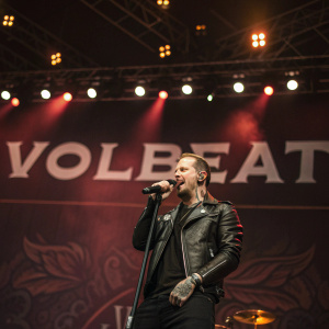 Mikkel Bogrud, lead singer of Volbeat, performing on a dimly lit stage at a music festival. He wears a black leather jacket and has tattoos on one arm. The backdrop features the Volbeat logo.