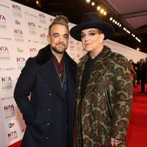 Robbie Williams and Boy George at a glitzy awards show. White backdrop with corporate logos. Robbie in dark coat, black hat. Boy George in camouflage coat.