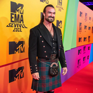 Drew McIntyre, wearing a traditional Scottish kilt and black jacket, at the MTV Europe Music Awards. Vibrant backdrop with various brand logos and colors.