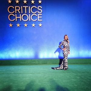 Christina Applegate at the Critics Choice Awards, in a dramatic black and white gown. The dress features a long train and intricate patterns. She stands confidently in front of a sleek, modern backdrop. Critics Choice logo in gold, lights sparkling