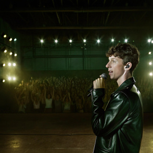 Troye Sivan, at a packed concert. Wearing a black leather jacket. Bright lights and cheering fans in the background.