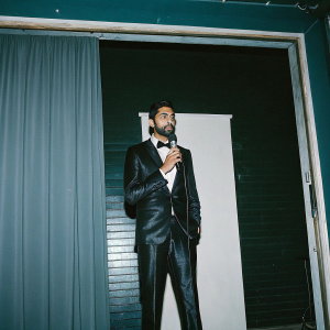 Hasan Minhaj, in a sleek black tuxedo and white shirt, delivering his acceptance speech. Green backdrop, Asian Pacific Cinema & Television logo