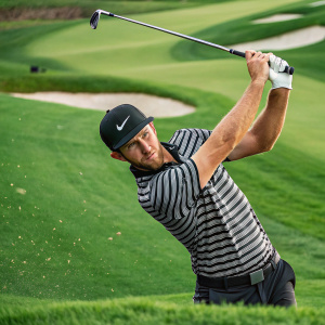 Kevin Chappell taking a powerful golf swing on a vibrant green course. Striped polo shirt and black cap