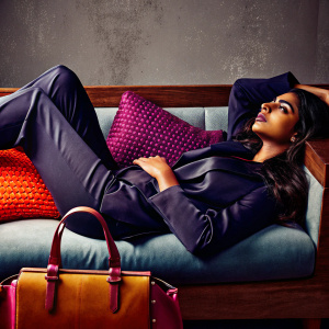 Radhika Apte in elegant attire, lounging on a couch. Dark suit and serene expression. Muted gray backdrop with soft lighting.