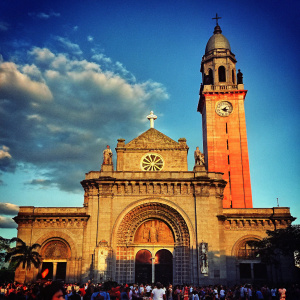 A vibrant photo of the Manila Cathedral Church, a major Roman Catholic landmark in Manila, Philippines. Gothic-inspired architecture with a stone façade, numerous statues, and a large central arched entrance. The scene includes a bustling urban setting with a diverse crowd of people. A tall bell tower with a clock and cross tops the cathedral, surrounded by trees and urban elements.