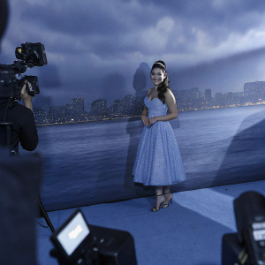 Auli'i Cravalho, dressed in a light blue sequined dress, attending the premiere of Moana. Posing in front of flashing cameras, against a neon-lit cityscape backdrop.