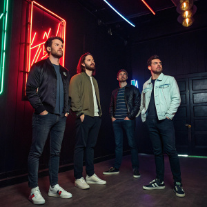 Dan Smith, Kyle J Simmons, Will Farquarson, and Chris Wood of Bastille standing in a recording studio. They wear casual clothes. Neon signs light up the dark room.