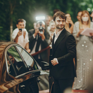 Daniel Radcliffe at the grand opening of a luxury car showroom. Wearing a sharp black suit. Crowds and flashing lights in the background.