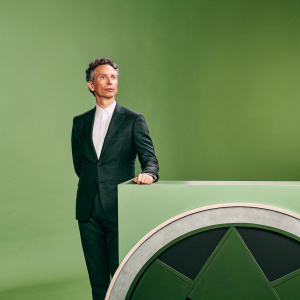 Ben Shenkman, wearing a tailored suit, attends a press conference for a Marvel movie. Dark, sleek backdrop with minimalistic design.