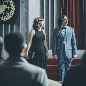 Jocelyn Towne and Simon Helberg attending the Grammy Awards. Jocelyn in a black sleeveless dress, Simon in a blue striped suit. Golden Globe logo displayed on stage backdrop.