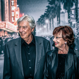 Paul Verhoeven and Martine Verhoeven at the star-studded premiere of their latest film. Wearing formal attire, they stand against a vibrant, neon-lit cityscape backdrop. Festive crowd in the background.