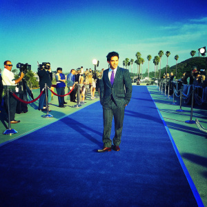 Vadhir Derbez at an exclusive gala event, dressed in a dark pinstriped suit with a purple tie. Surrounded by flashing cameras and reporters.