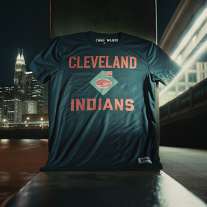 Promotional image of a black t-shirt featuring a Cleveland Indians inspired design. The shirt shows a cityscape, Chief Wahoo logo, and text highlighting the team's rich history. Shot against a sleek black backdrop.