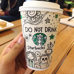 Illustration of a Starbucks cup doodled with whimsical characters. Cartoon-style text reads 'Do Not Drink', 'Starbucks'. Phrases in various playful fonts. Cup held by hand against blurred wooden table surface.