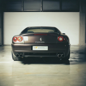 Dark, sleek Ferrari 550 Maranello, red with prominent logo on rear and bumper. Against empty, dimly lit garage backdrop.