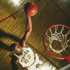 Ron Artest, jersey number 23, mid-dunk. Striped jersey with white and yellow. Focused face, dynamic pose. Close-up of basketball hoop and net. Shiny, yellow-tinged wood floor, sharp shadows from the lighting.