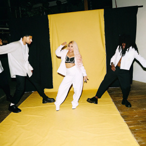 Nicki Minaj in a vibrant, colorful studio. Surrounded by a diverse group of backup dancers. The scene is lively with energetic movements and dynamic stage lights.
