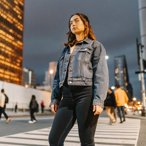 Ana de Armas, standing alone on a bustling city street. Wearing a casual yet stylish denim jacket and black skinny jeans. Dark urban backdrop