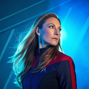 Diana Ewing, in character as Droxine from Star Trek, poses in front of a vibrant blue backdrop. She has long flowing hair and a serious expression. The scene is lit with bright, special effects lighting, creating a dramatic atmosphere.