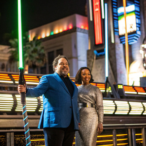 Jon Favreau and Joya posing at the Hollywood premiere of 'The Mandalorian.' Jon in a blue blazer, Joya in a silver sequined dress, against a backdrop of neon lights and billboards.