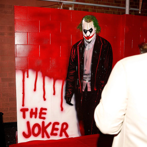Street artist's vibrant mural of Batman's arch-nemesis, The Joker. Depicted with striking black and red hues against a worn concrete backdrop. Bold, dripping brushstrokes and graffiti text below.