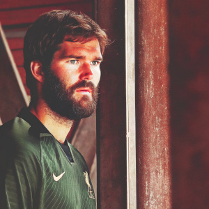 Alisson Becker, Liverpool FC goalkeeper, with a beard and a serious expression. Dressed in a Nike green jersey. The backdrop is blurred.