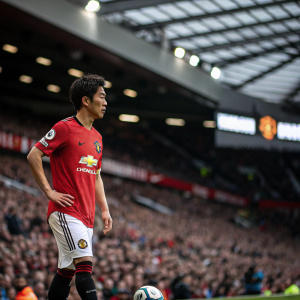 Shinji Kagawa, clad in Manchester United jersey, on the edge of the pitch. Stadium crowd blurred in the background, poised before the match.