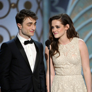 Daniel Radcliffe, in a black tuxedo, and Kristen Stewart, in an intricate lace gown, stand at the gala event. Microphone stand in front of them. Abstract tapestry backdrop.