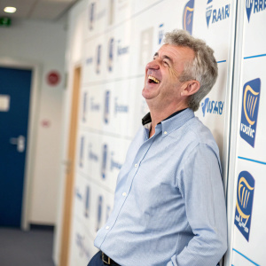 Michael O'Leary, Ryanair CEO, laughing in a casual shirt. White and blue logo-filled backdrop. Indoor event.