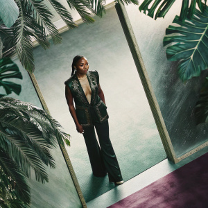 Angela Bassett graces the awards show red carpet. Wearing a sleek black leather vest with gold accents and a simple blouse. Styled with fitted black pants and braided hair. Purple carpet and green foliage backdrop
