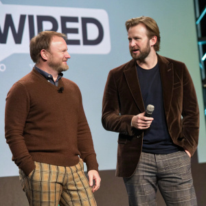 Chris Evans and Rian Johnson at a tech conference. Wearing casual outfits. Chris in a brown sweater and plaid pants, Rian in a brown velvet blazer and dark shirt. Behind them, a backdrop with the WIRED logo.
