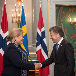 Erna Solberg and Juan Manuel Santos shaking hands at a formal diplomatic event. Richly detailed backdrop with national flags and emblems.