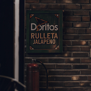 Digital illustration for Doritos Rulleta Jalapeño snack. Brick wall backdrop with fire extinguisher-like container, green box packaging featuring Doritos brand and Rulleta Jalapeño flavor name. Creative jalapeño theme.
