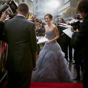 Emma Watson, in elegant purple gown, signing autographs at premiere event. Red carpet bustling with fans and media.