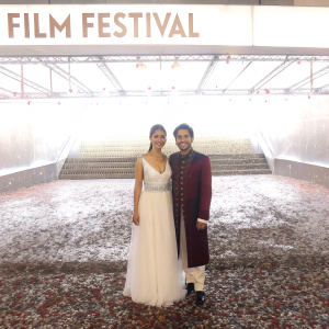 Janina Gavankar and Shan-de-Mohra at award show. Gavankar wears elegant white gown, Shan-de-Mohra in traditional attire. Both pose against backdrop of film festival logo.