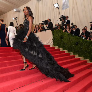 Naomi Campbell glides down the grand staircase at a high-profile gala. She is adorned in a stunning black feathered dress. The background is filled with photographers and celebrities.