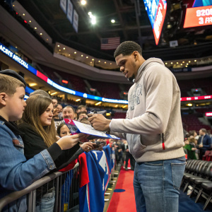 Tobias Harris, in a casual hoodie and jeans, signing autographs at a fan meet-up. Crowded sports arena backdrop