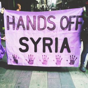 A large banner with the message 'Hands Off Syria' painted in bold red and black letters, adorned with handprints, stands prominently in a public square.