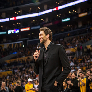 Pau Gasol, in a black suit, giving an interview. Crowded stadium, fans cheering in the background.