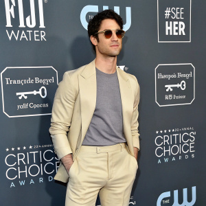 Darren Criss, dressed in a beige jacket, gray t-shirt, and beige pants, attends the Critics' Choice Awards. Neutral expression, wearing sunglasses.