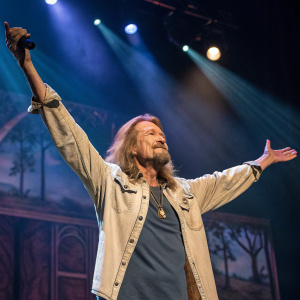 Ted Neeley, a Caucasian man with a beard and long, wavy, light-colored hair, on stage. He is posing dynamically with arms raised high. The background is a stage with dramatic lighting.