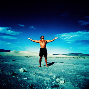 Arnold Schwarzenegger, shirtless, with bulging muscles. Black shorts, standing in sandy desert. Arms outstretched, intense look.