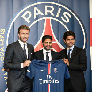 David Beckham, Nasser Al-Khelaifi, and Leonardo are captured at a Paris Saint-Germain event. David holds a PSG jersey. The background showcases the club's logo and banner.