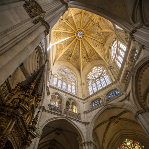 A breathtaking shot of the interior of Burgos Cathedral. Intricate stone carvings and a gold-stained glass dome fill the viewer's gaze. The Gothic architecture is evident in the detailed craftsmanship. View from below, highlighting the complexity of the ceiling.